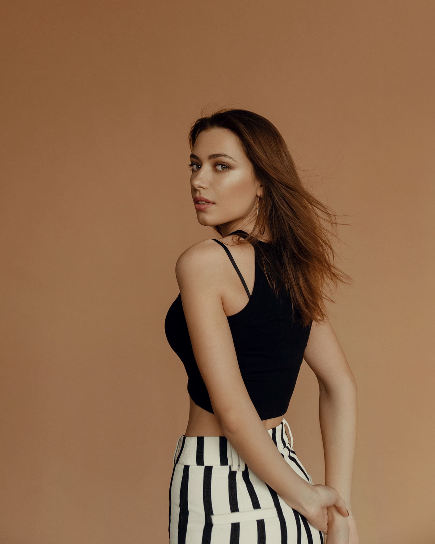 Mujer de cabello castaño de perfil ante fondo beige, top negro y pantalón a rayas verticales, manos entrelazadas tras la espalda.