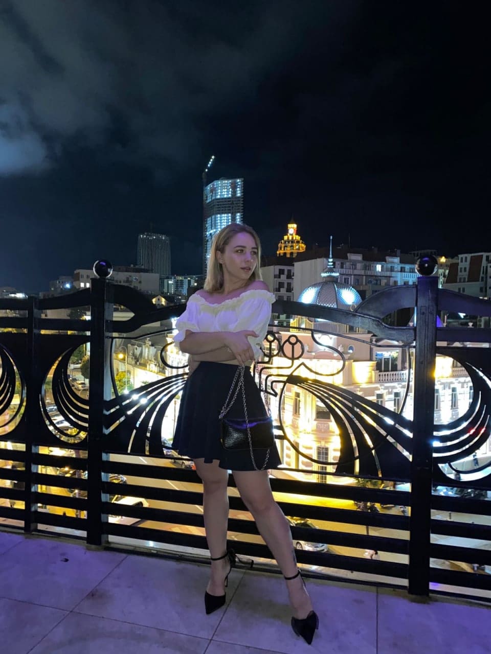 Mujer rubia en balcón nocturno con blusa blanca, falda negra y tacones, frente a barandilla y ciudad iluminada.