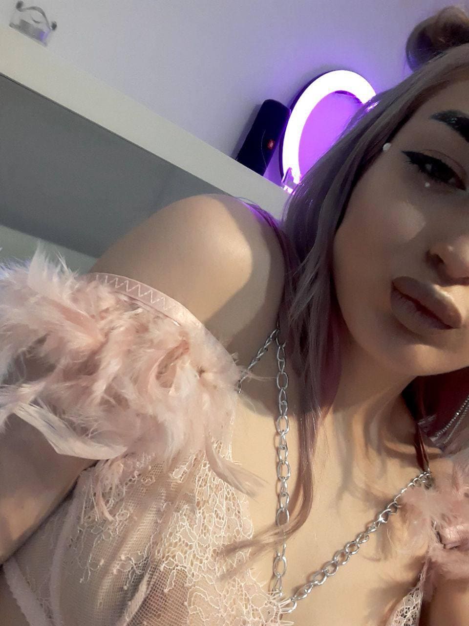 Selfie cercana de persona con cabello rosa pálido usando top de encaje transparente con plumas y collar plateado.