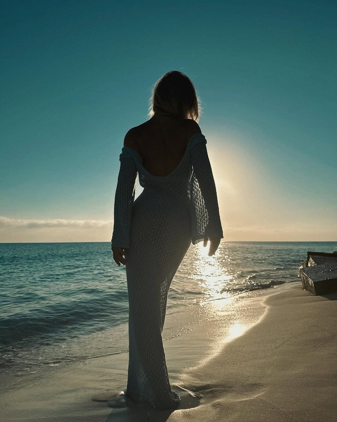 Persona de pelo largo de espaldas al mar al atardecer, con vestido largo de punto abierto que descubre hombros y espalda en la arena.