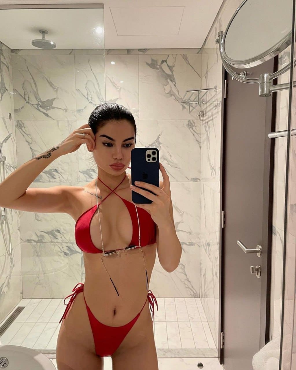 A woman takes a mirror selfie in a marble bathroom wearing a red string bikini, one hand raised to her dark hair.
