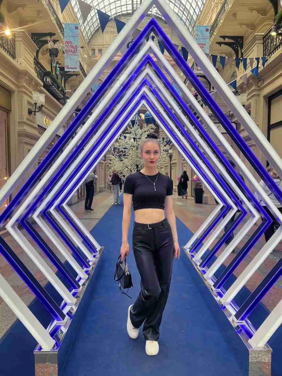 A woman walks down a blue carpet through illuminated arches, wearing a black crop top, leather pants, and white sneakers.
