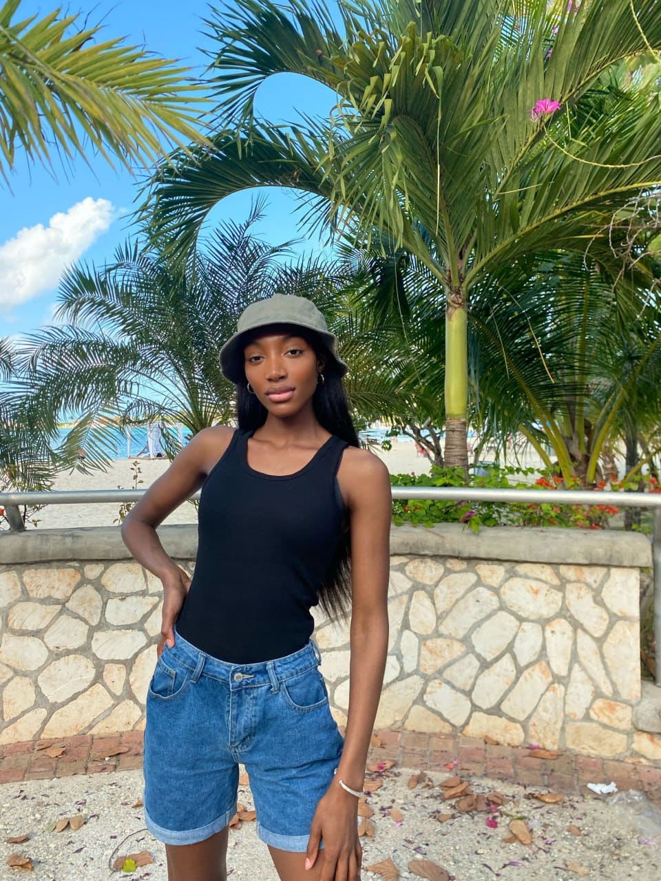Donna all'aperto davanti a un muro di pietra con cappello grigio, top nero e pantaloncini in denim, mano sulla vita.
