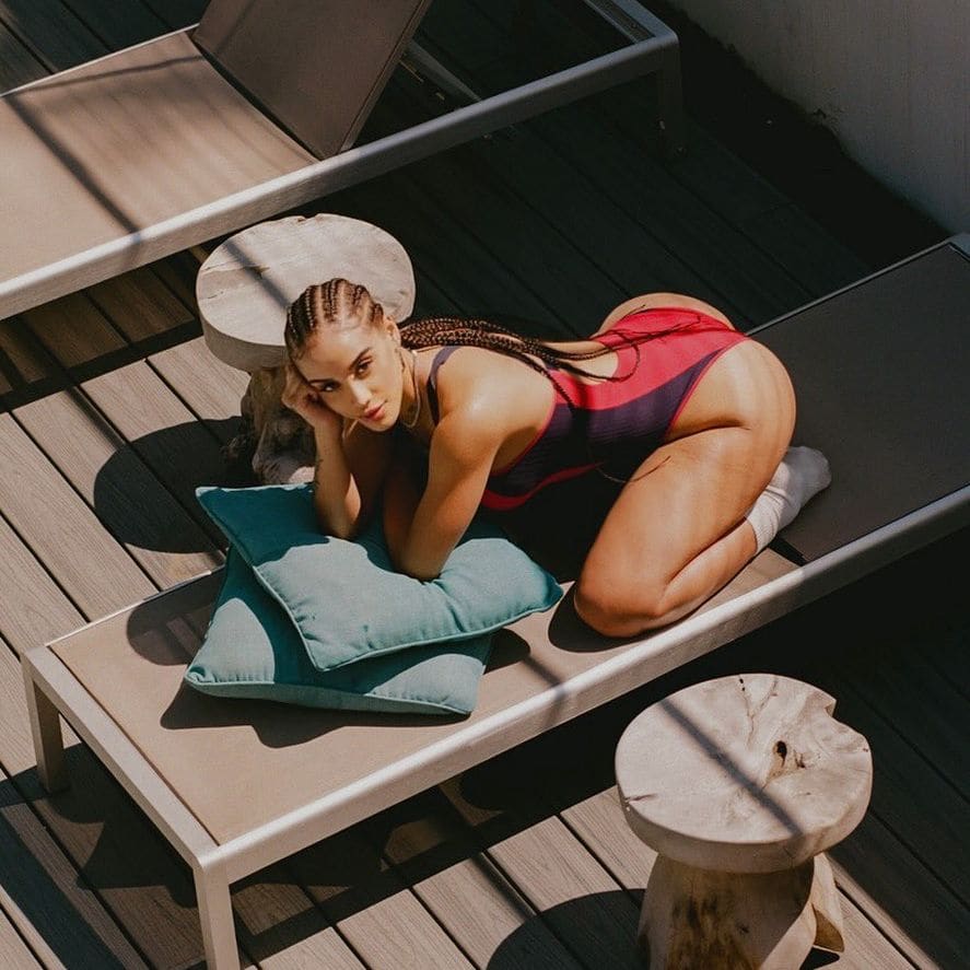 Mujer con trenzas arrodillada en una tumbona al aire libre, con bañador rojo y negro y calcetines blancos.
