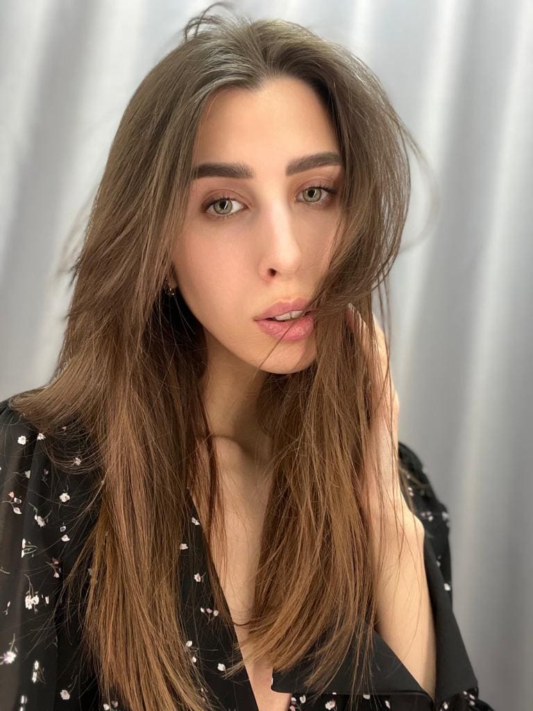 Primer plano de mujer tocándose el cabello, viste top negro estampado floral con escote V profundo ante fondo gris claro.