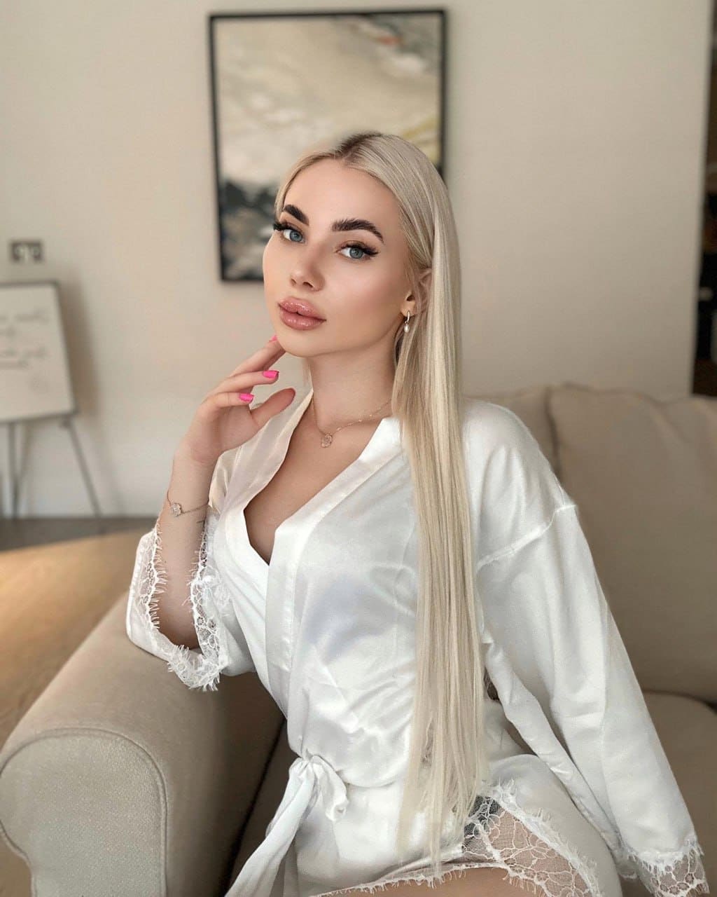 Diana sits on a beige sofa in a white satin robe with lace trim, hand to chin, before an abstract painting.