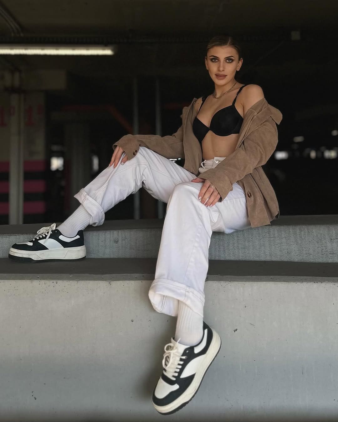 Mujer de cabello oscuro sentada en un banco interior con poca luz, con sujetador negro y jeans blancos.