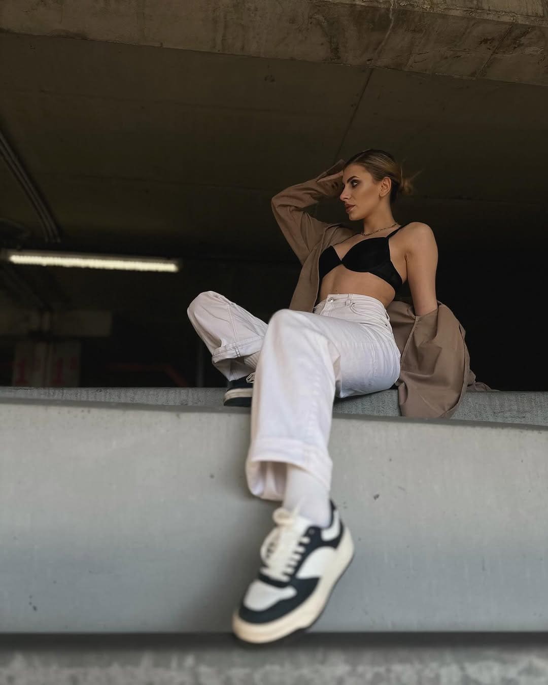 Mujer rubia sentada en una acera bajo un paso inferior, con sujetador negro y chaqueta sobre hombros.