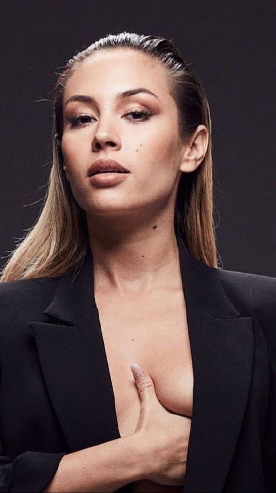 Mujer con blazer negro de abertura profunda posa con la mano derecha sobre el pecho, contra un fondo de estudio oscuro.