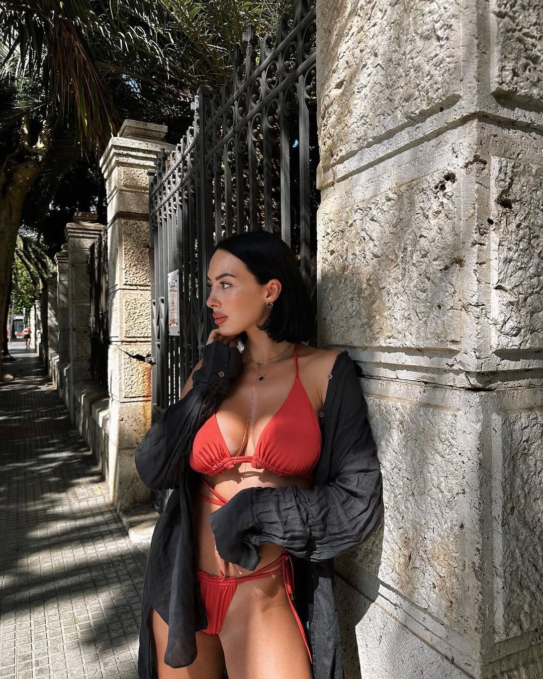 Mujer de bikini rojo y camisa negra abierta apoyada en muro de piedra junto a reja.