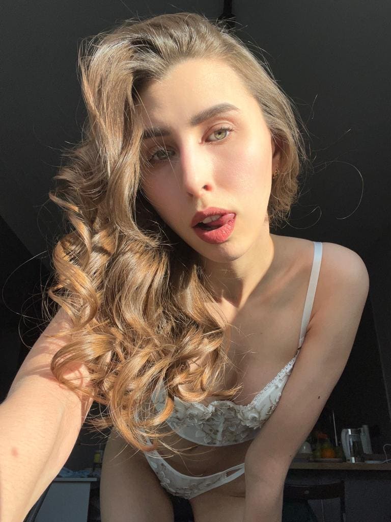 Mujer rubia de cabello ondulado se inclina hacia adelante vistiendo lencería blanca de encaje contra un fondo oscuro.