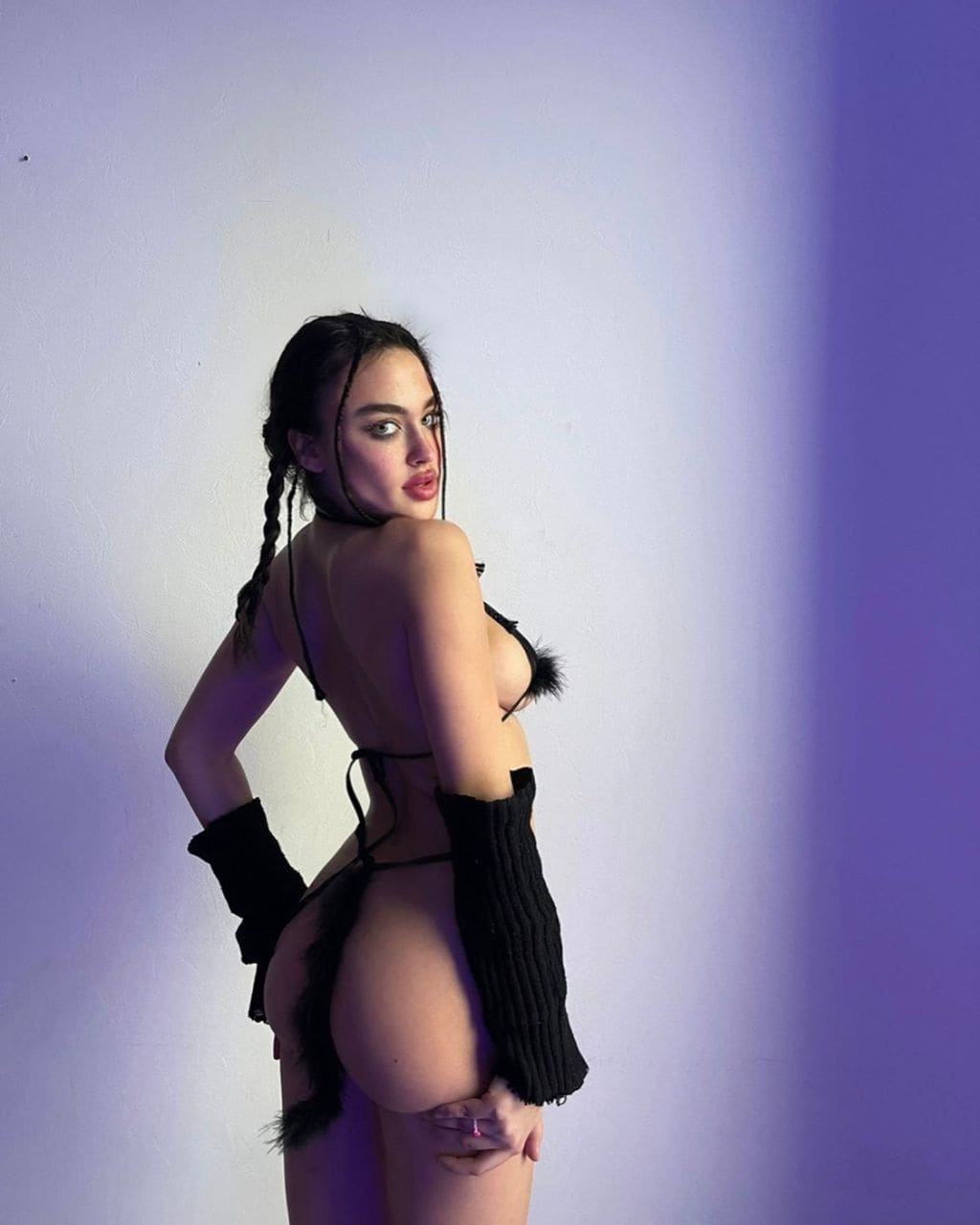 Mujer de perfil con trenzas frente a pared lisa, bikini negro de plumas, manga tejida negra y accesorio de cadera, mano en la cintura.