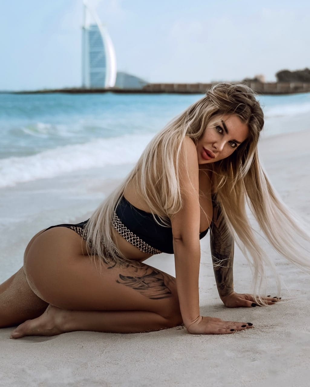 Mujer rubia arrodillada en la playa con bikini negro y cinturón de cadena, mirando a cámara frente al océano.