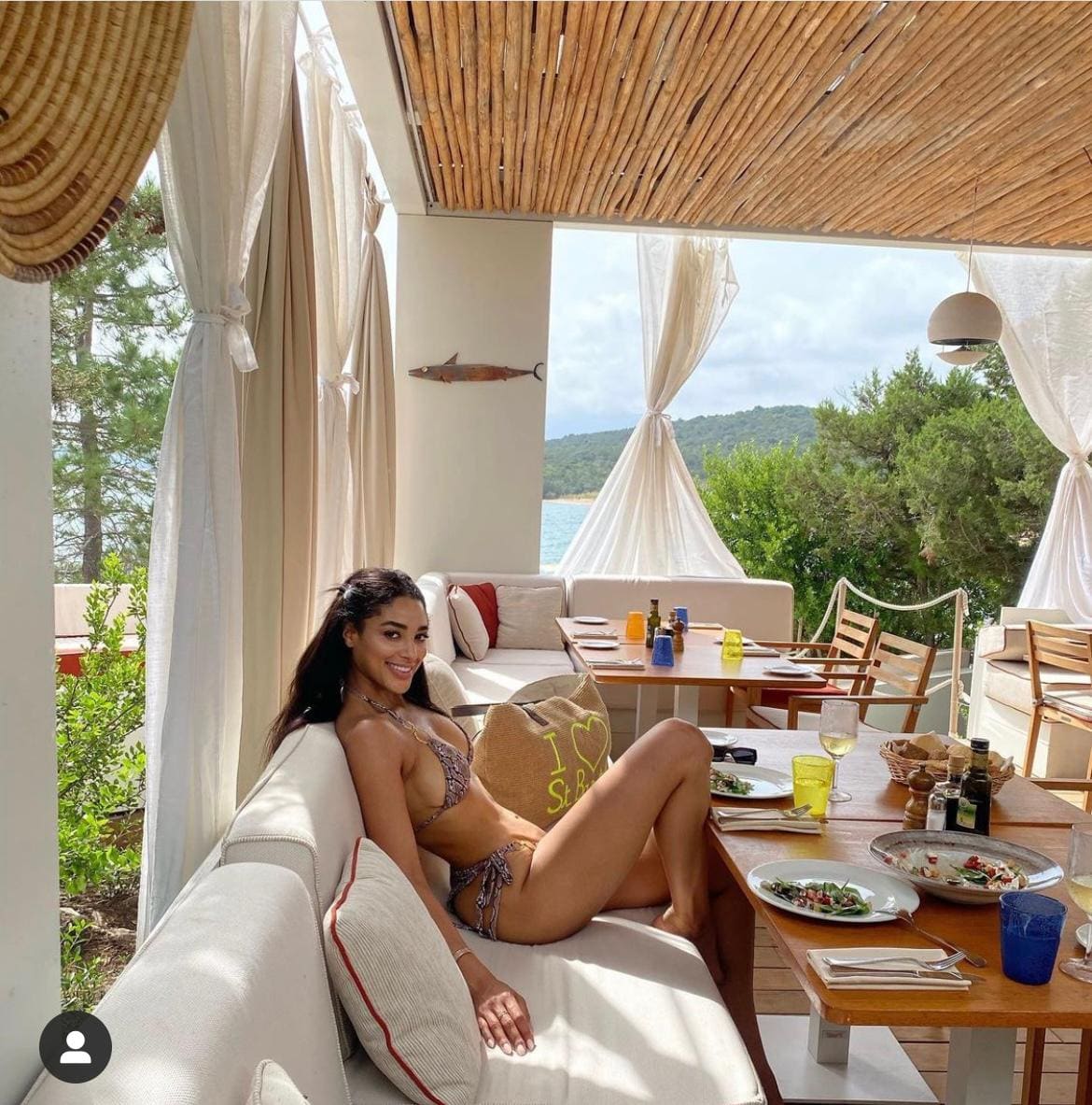 Donna in bikini fantasia sdraiata su divano bianco all'aperto, gambe incrociate, con vista su alberi e acqua.