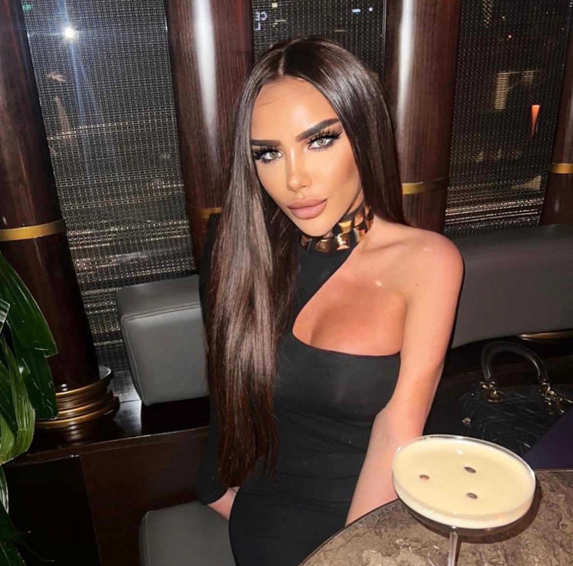 A woman sits at a dimly lit table in a black one-shoulder dress and patterned choker, with a cocktail glass in the foreground.