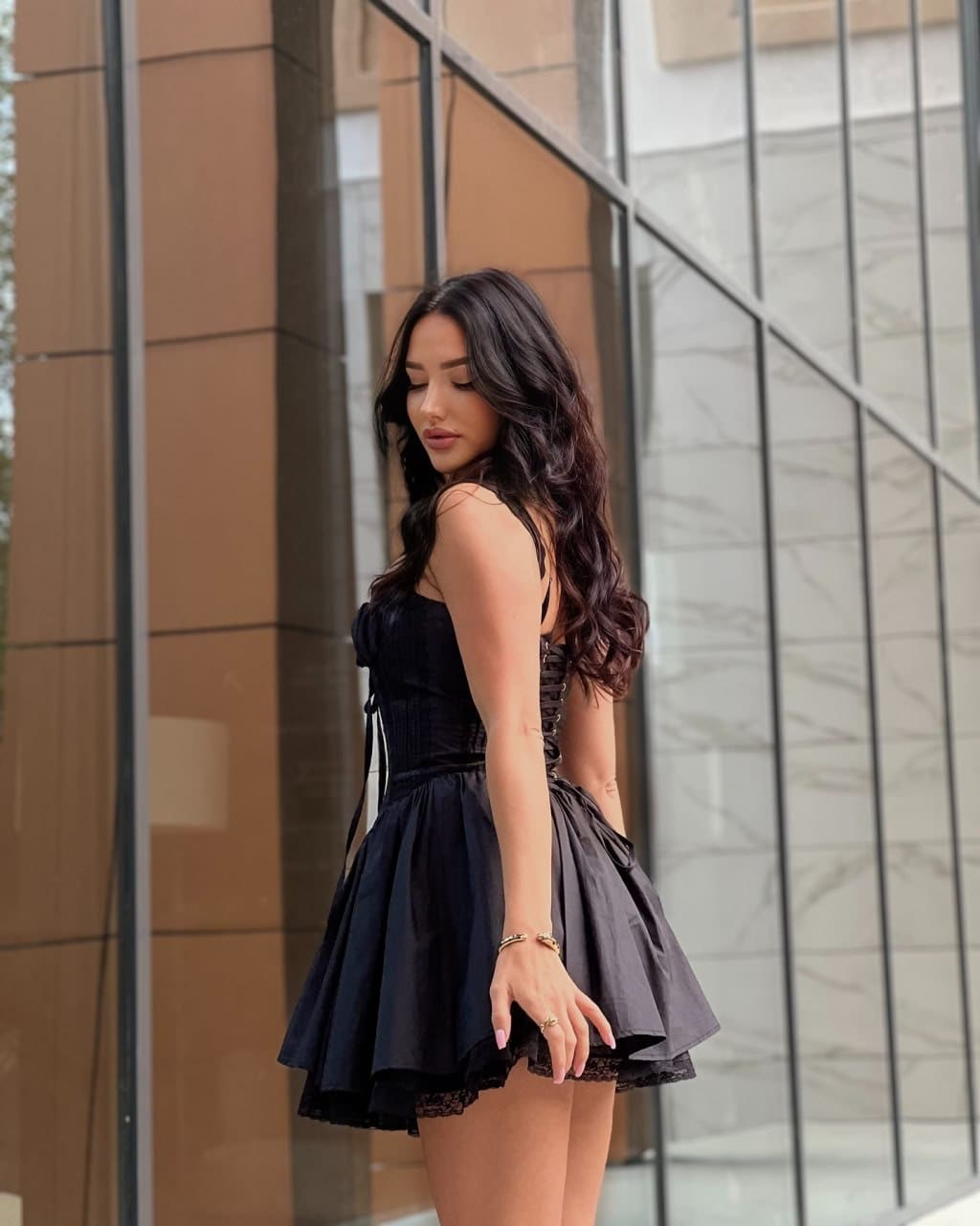 De pie ante un edificio con cristal, viste un vestido negro corto sin mangas y mira hacia abajo.