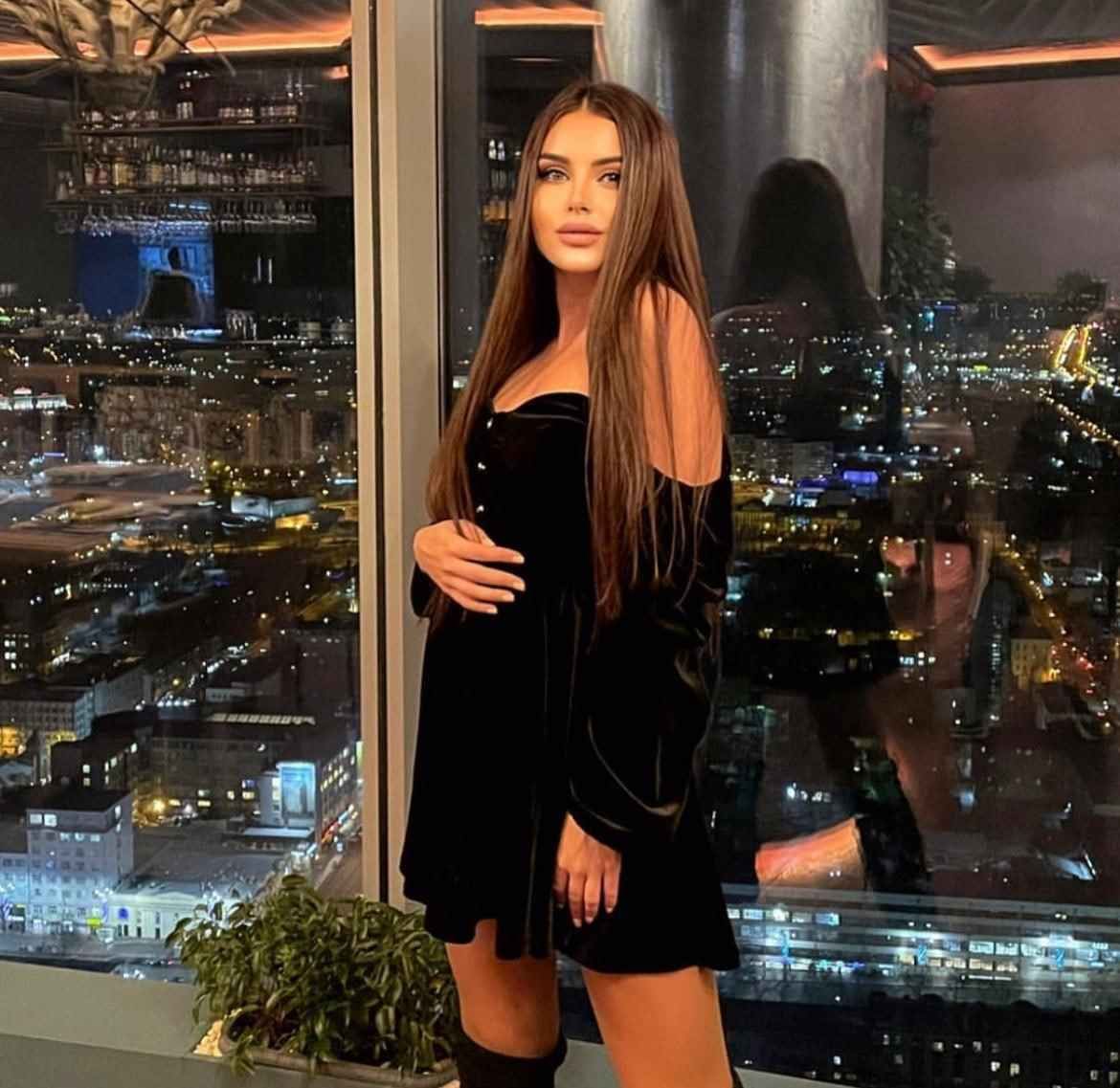 A woman in a short black velvet dress stands by a window overlooking a city at night, her right hand resting on her hip.