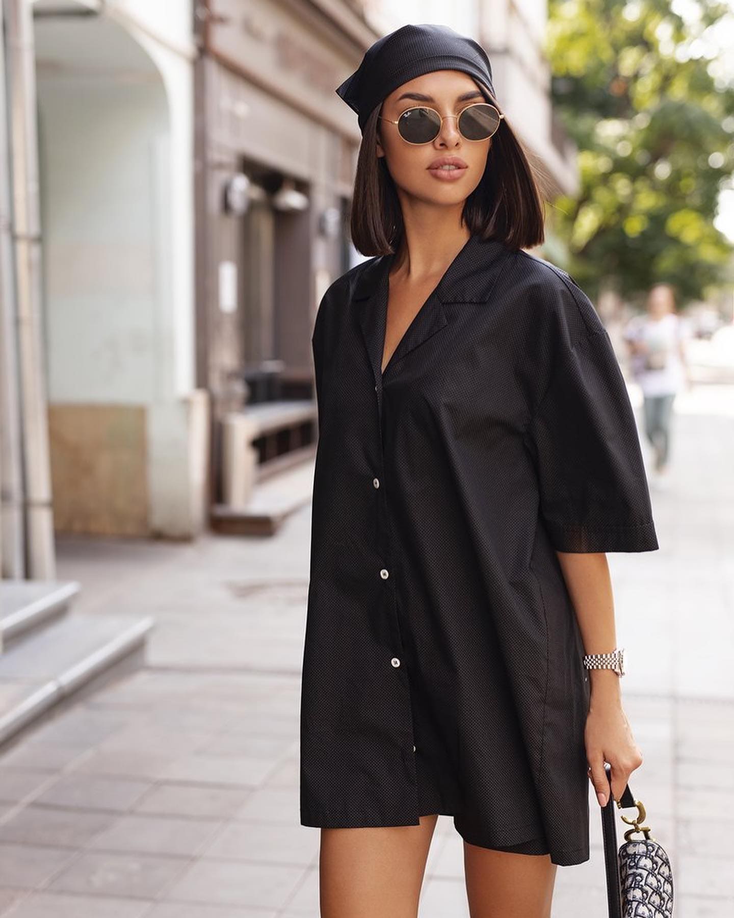 Mujer con cabello oscuro hasta los hombros en una acera urbana, vestida con un vestido-camisa negro, tocado, gafas redondas y reloj, sosteniendo un bolso estampado.