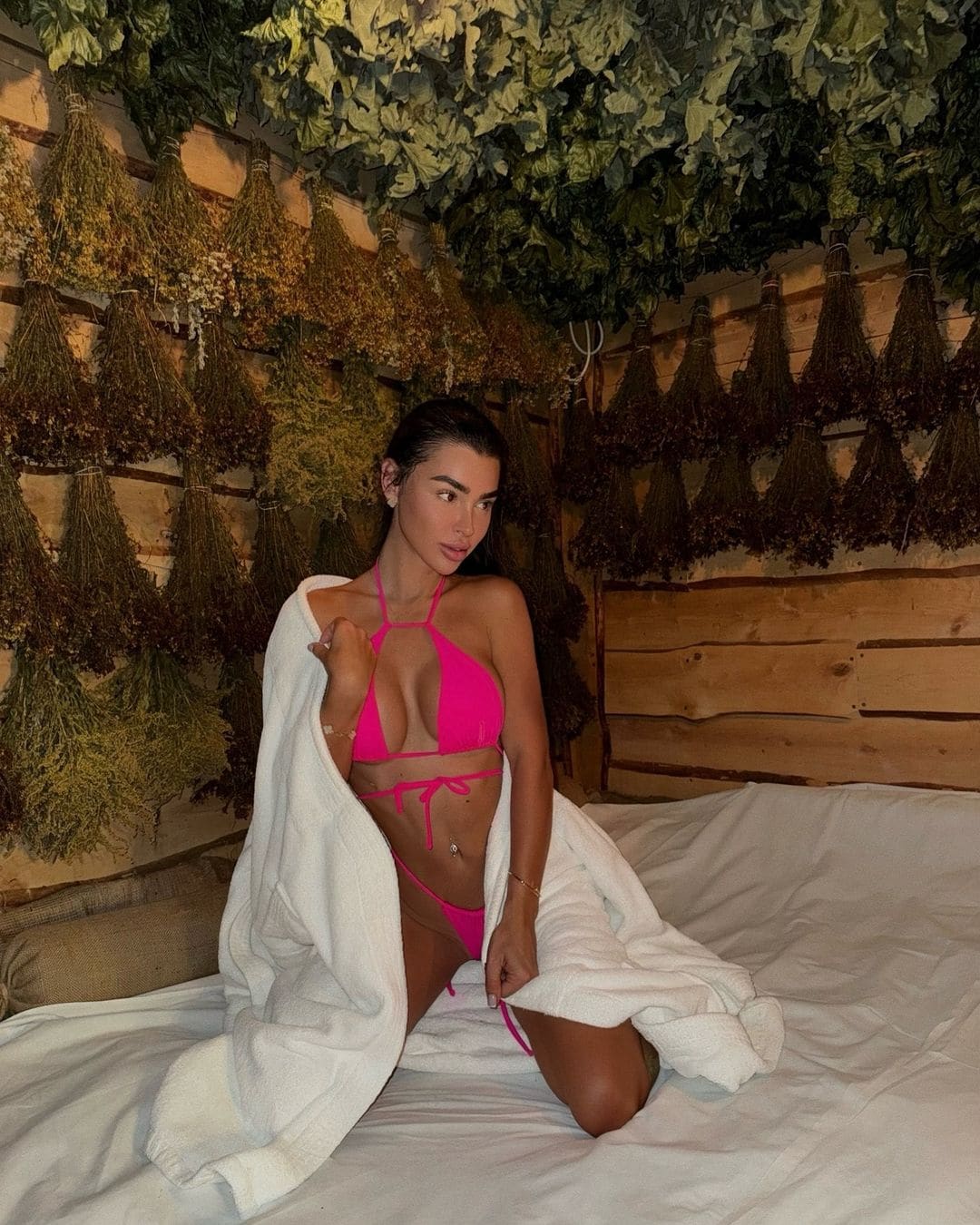 Mujer con el cabello recogido sentada en cama blanca, con bikini rosa brillante y toalla blanca sobre los hombros, frente a pared de madera.