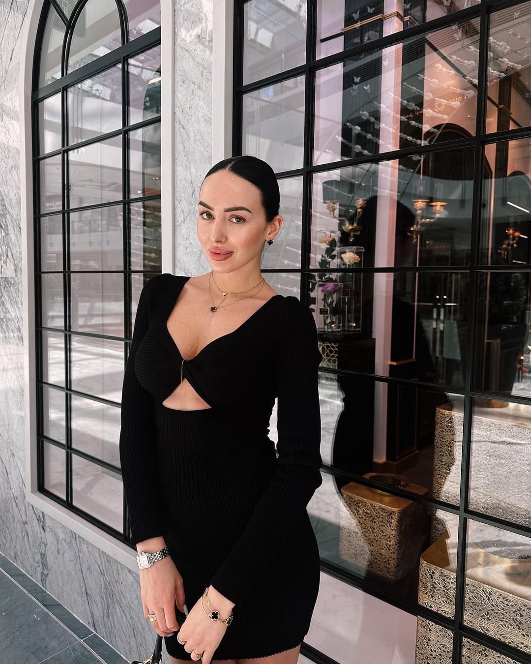 Mujer de vestido negro ceñido con corte en el abdomen ante gran ventana de marco negro.