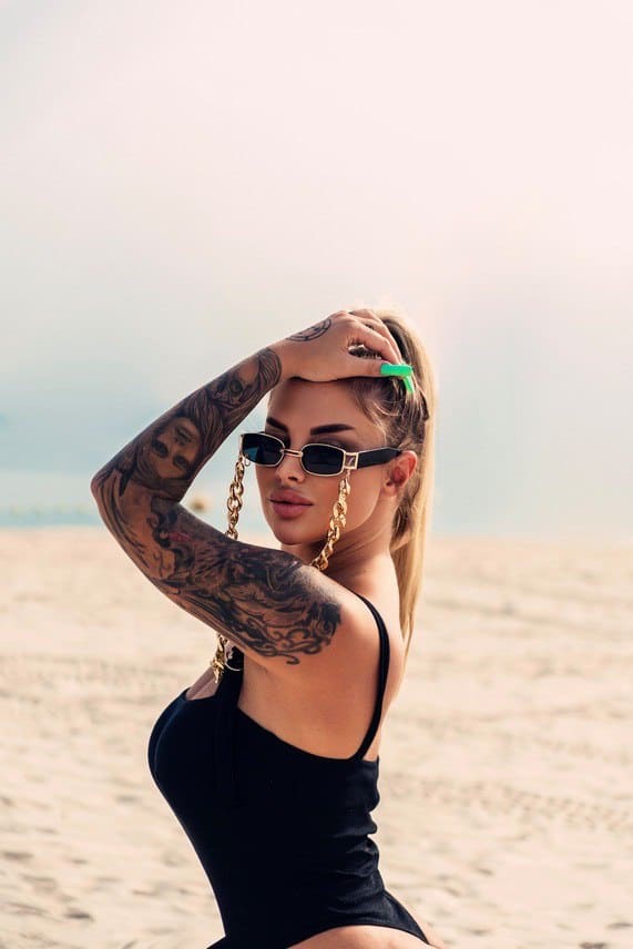 Mila sits on a sandy beach in a black one-piece swimsuit and gold-chained sunglasses, holding her hand to her head.