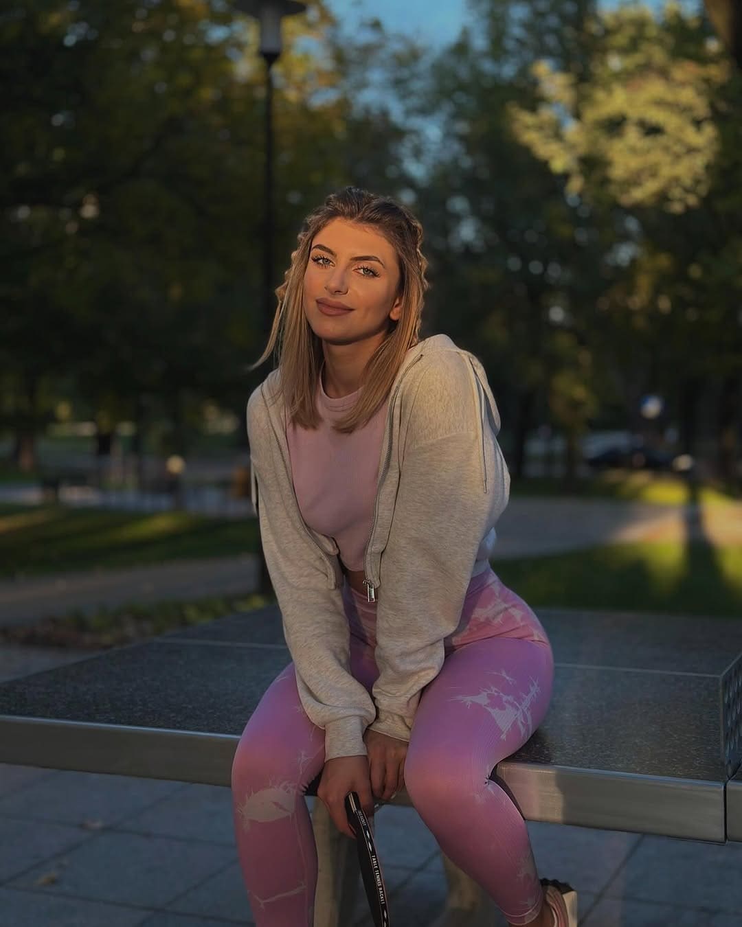 Mujer rubia sentada en un banco de piedra en un parque, con sudadera gris y leggings rosas.