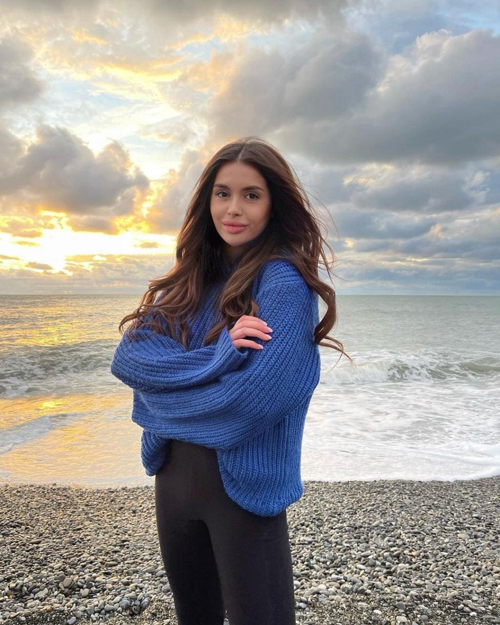 Mujer con cabello castaño ondulado de pie en una playa de grava con los brazos cruzados, luciendo un suéter azul grueso de punto y leggings negros, con el océano y el atardecer nublado al fondo.