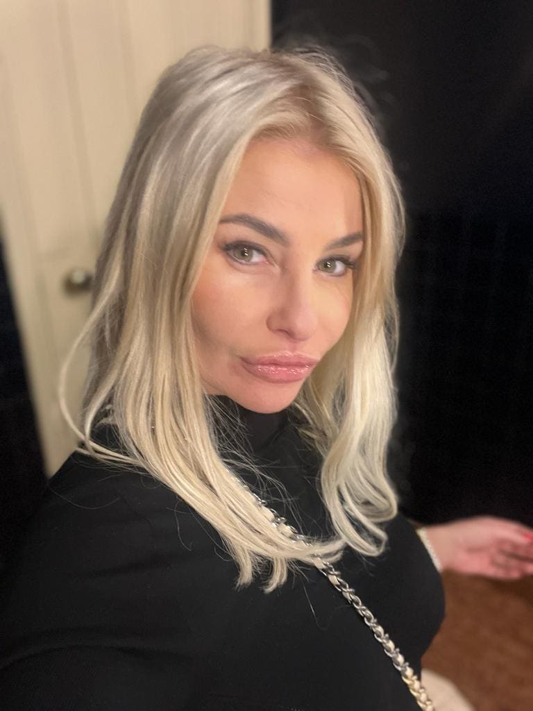 Mujer rubia platino con cuello alto negro y cadena plateada, posando frente a una puerta.