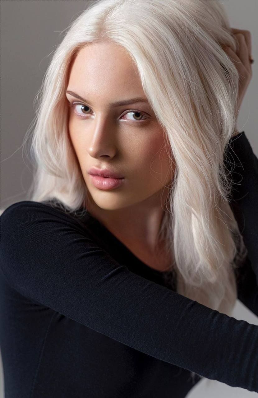 A woman in a black long-sleeved top poses against a gray wall, hand behind her head.