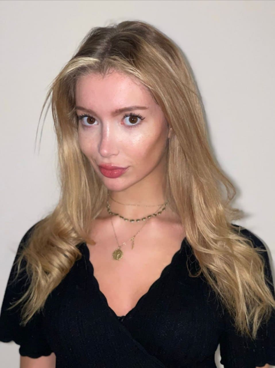 Clara standing in a black V-neck top with layered necklaces against a wall.