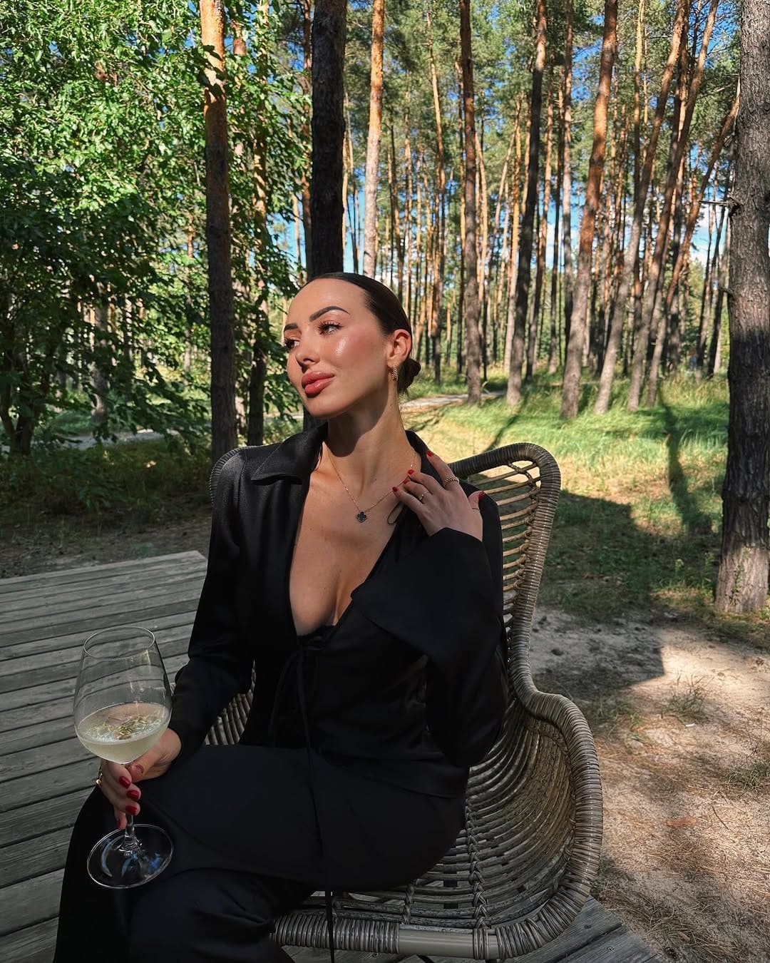 Mujer de vestido negro escotado sentada en silla tejida entre pinos con luz natural.