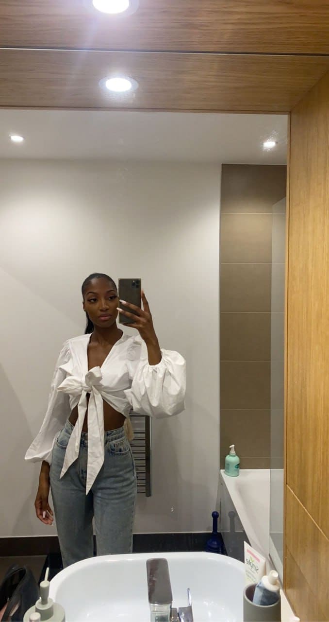 Donna con camicia bianca annodata in vita e jeans chiari, selfie allo specchio in bagno con lavandino e articoli visibili.