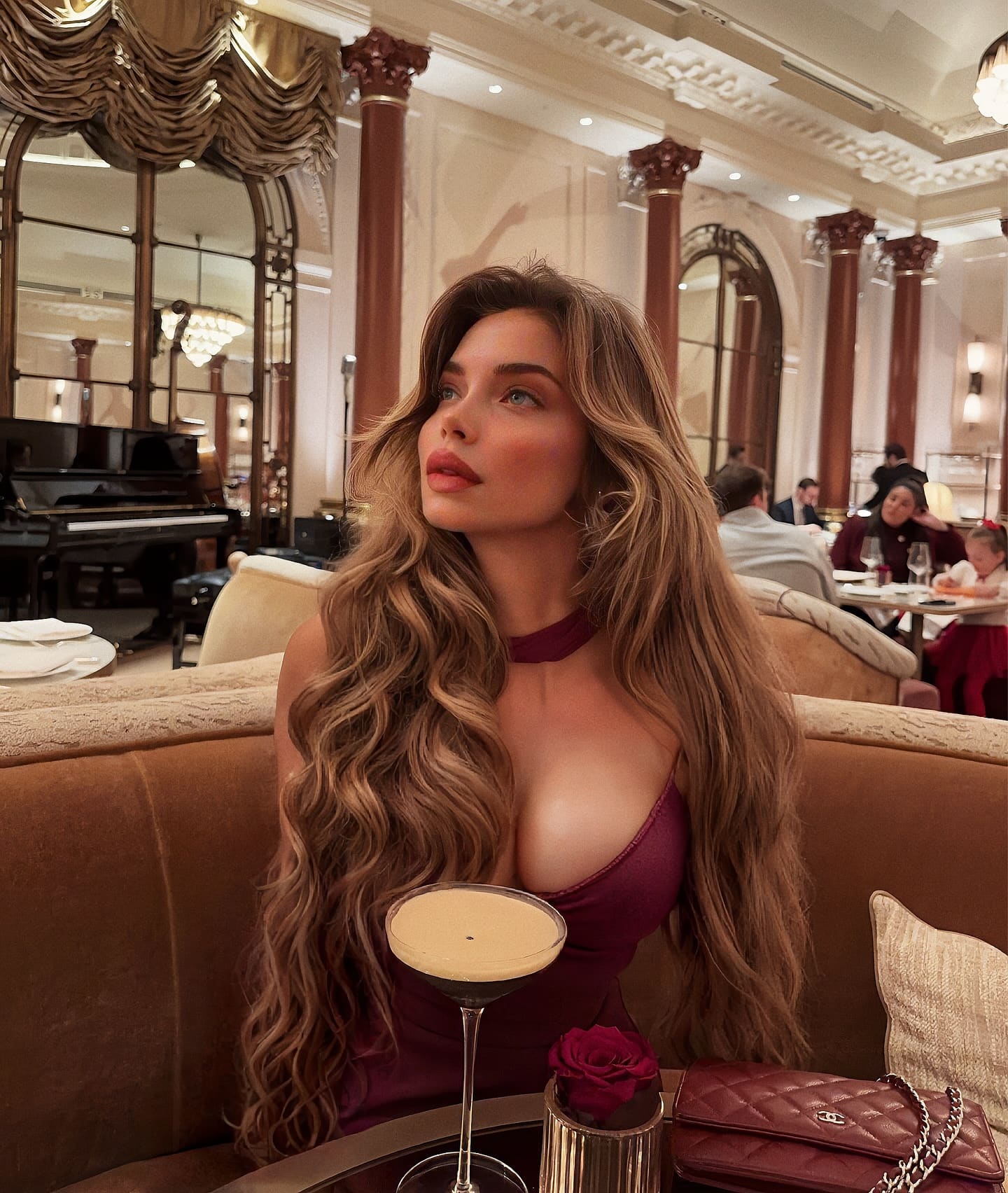 Alenia, con cabello castaño ondulado, sentada en un sofá de una sala ornamental, vestida con un vestido sin mangas color vino y un collar a juego.