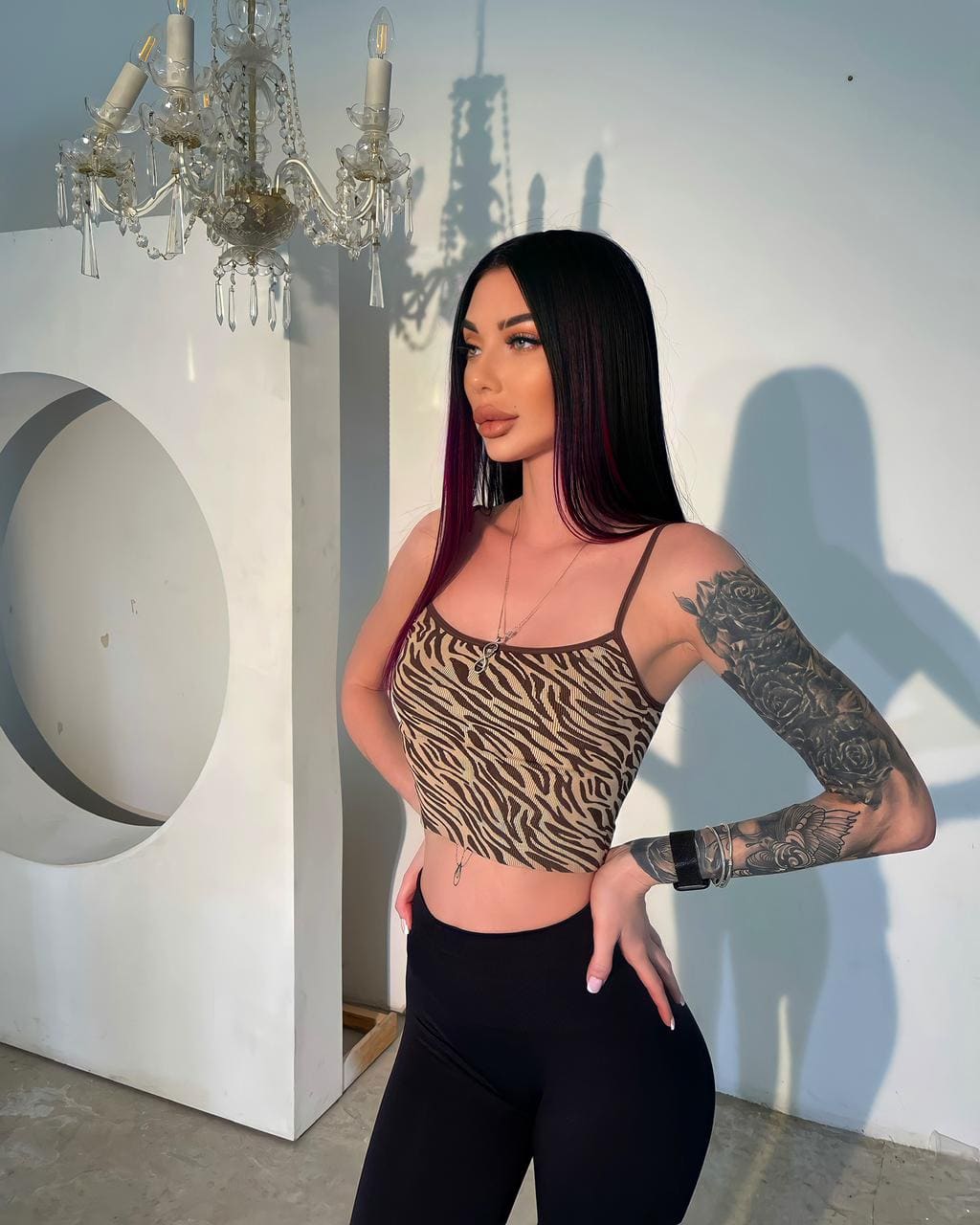 Melissa con cabello oscuro y mechas rojas, top estampado cebra y leggings negros, de pie en sala con candelabro.