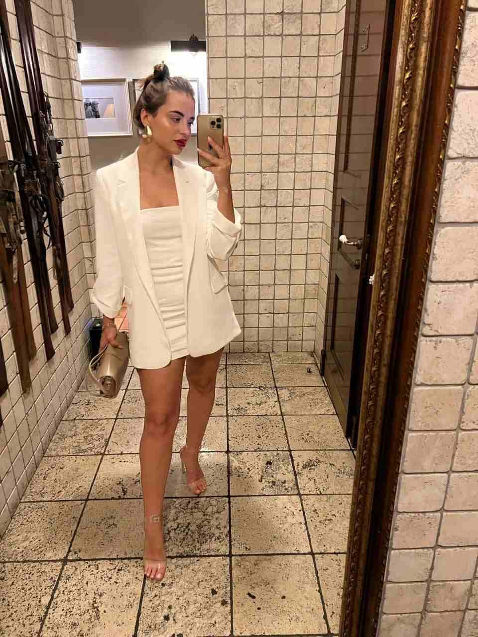 Donna con chignon che si fotografa allo specchio in corridoio piastrellato, giacca bianca su minidress bianco, tacchi trasparenti e clutch metallico.