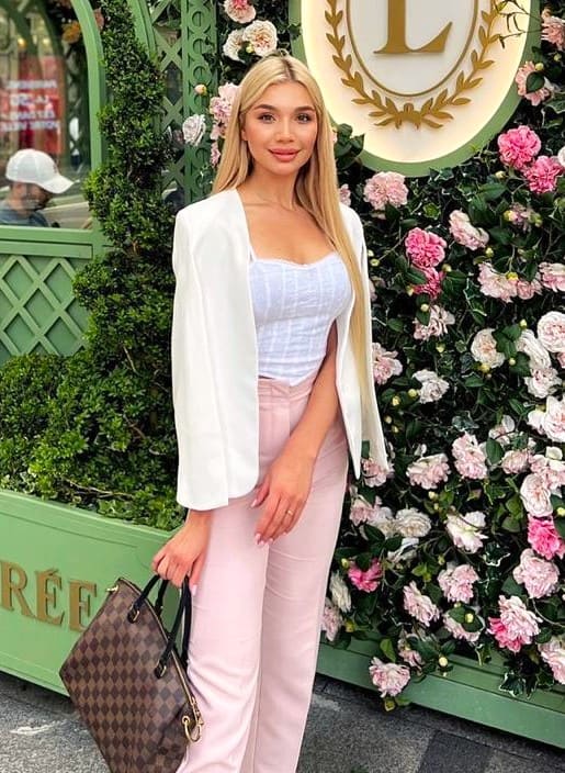 Alisson in a white blazer and pink trousers holds a handbag before a floral wall.