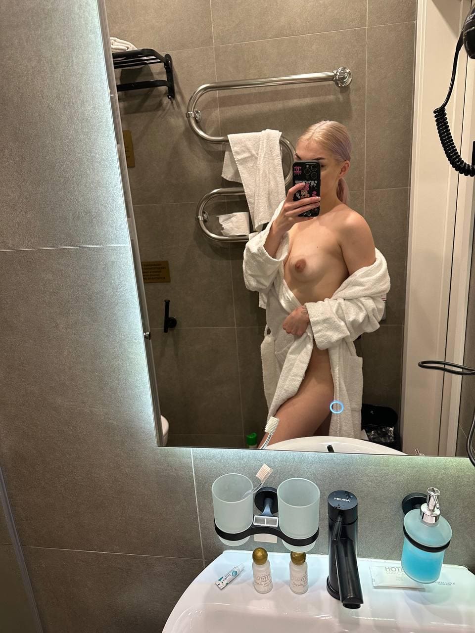 Persona de cabello claro en baño de azulejos sosteniendo un teléfono para selfie, con bata blanca abierta sobre torso desnudo.
