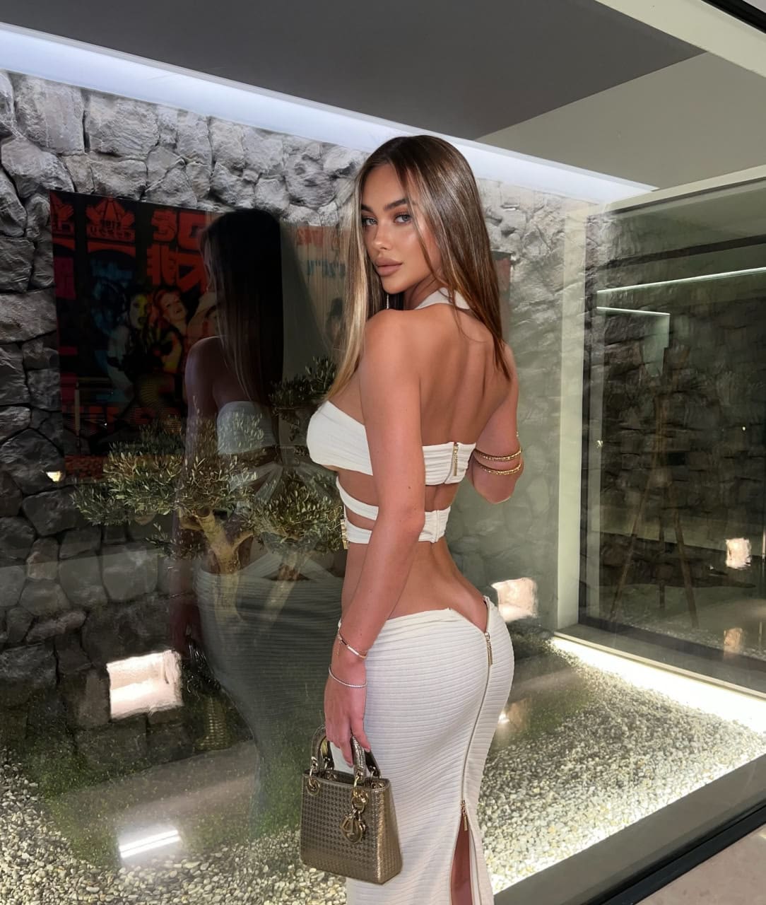 Mujer de perfil junto a una pared de cristal, vestida con conjunto blanco sin tirantes y falda aberturas, sosteniendo un bolso pequeño.