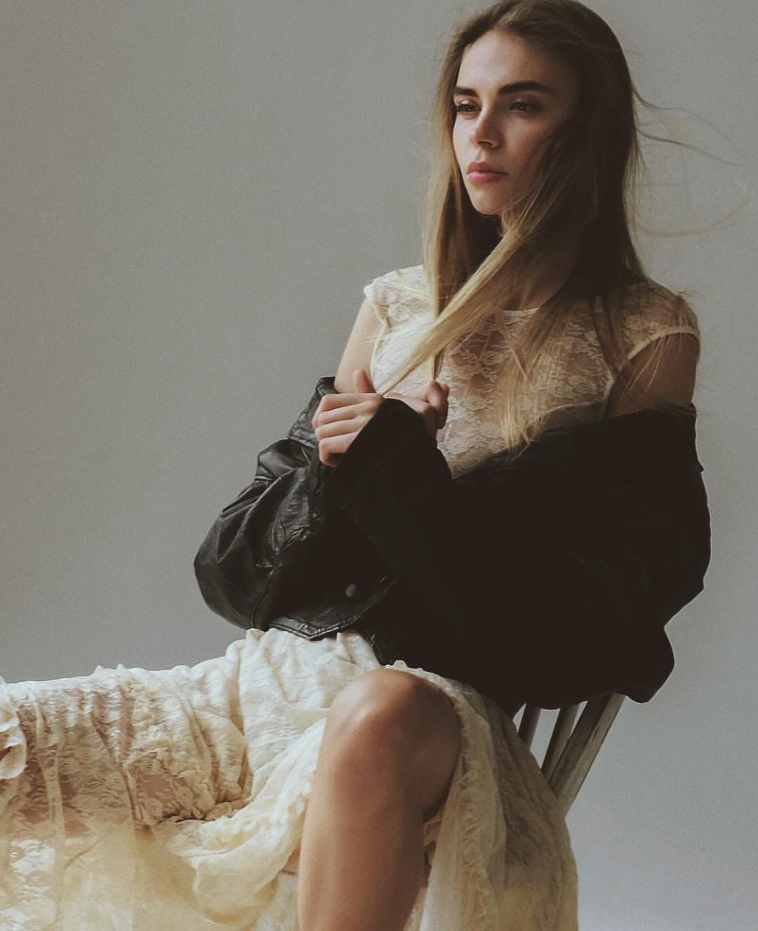Joven rubia sentada en silla de madera, vestido blanco de encaje transparente y chaqueta de cuero negra sobre las rodillas.