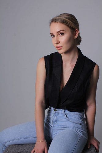 A woman sits facing the camera in a black V-neck wrap top and light blue jeans, arms at her sides on a gray backdrop.