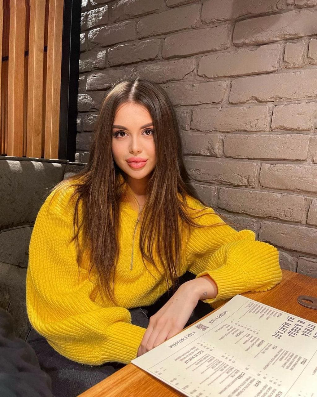 Kayla con cabello castaño largo sentada en una mesa de madera frente a una pared de ladrillo gris, vistiendo un suéter amarillo ribeteado y apoyando los brazos sobre un menú impreso.