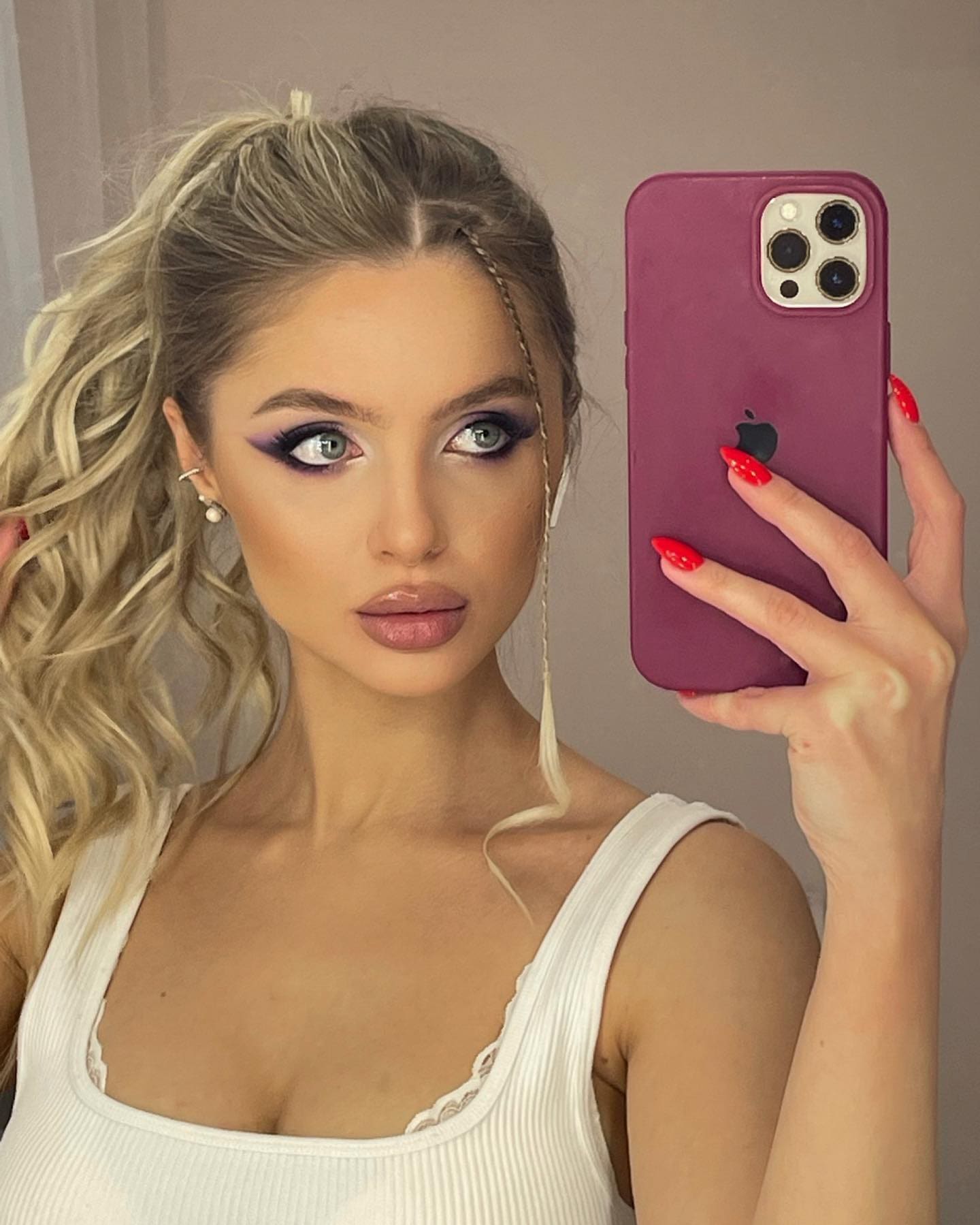 Mujer rubia con coleta media sujetando un móvil morado ante el espejo, vestida con top blanco de tirantes.
