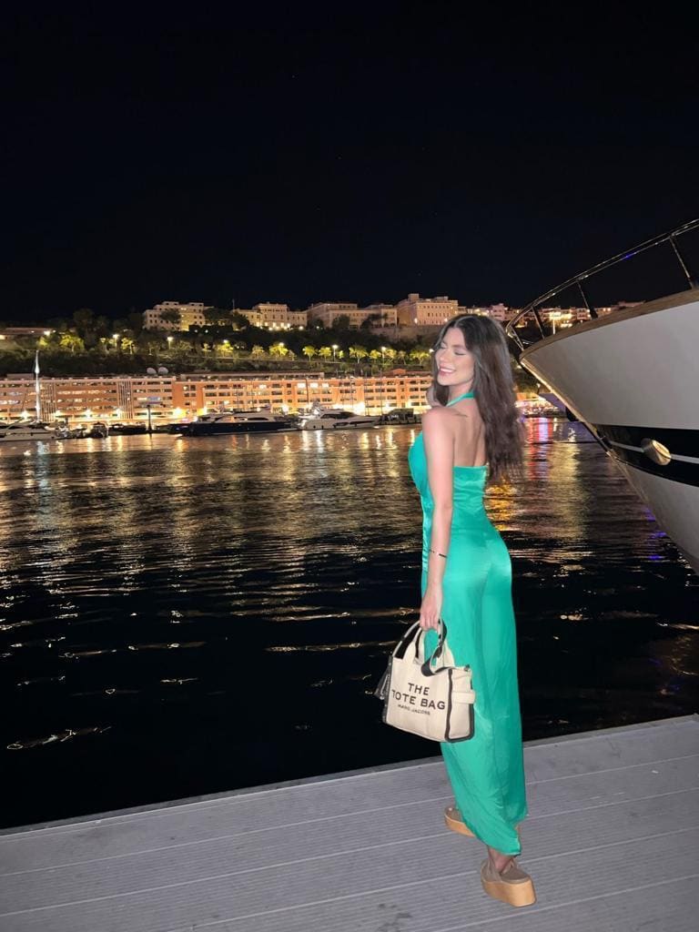 De pie en muelle nocturno con vestido verde largo, sandalias y puerto iluminado.