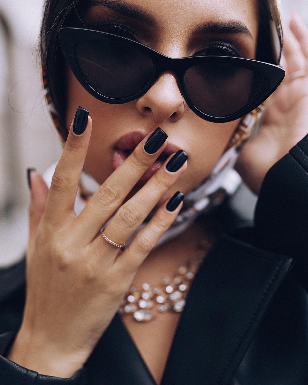 Primer plano de una mujer con el cabello recogido, usando gafas de sol negras de ojos de gato, blazer negro, bufanda estampada y collar de cuentas gruesas, con la mano cerca de los labios.
