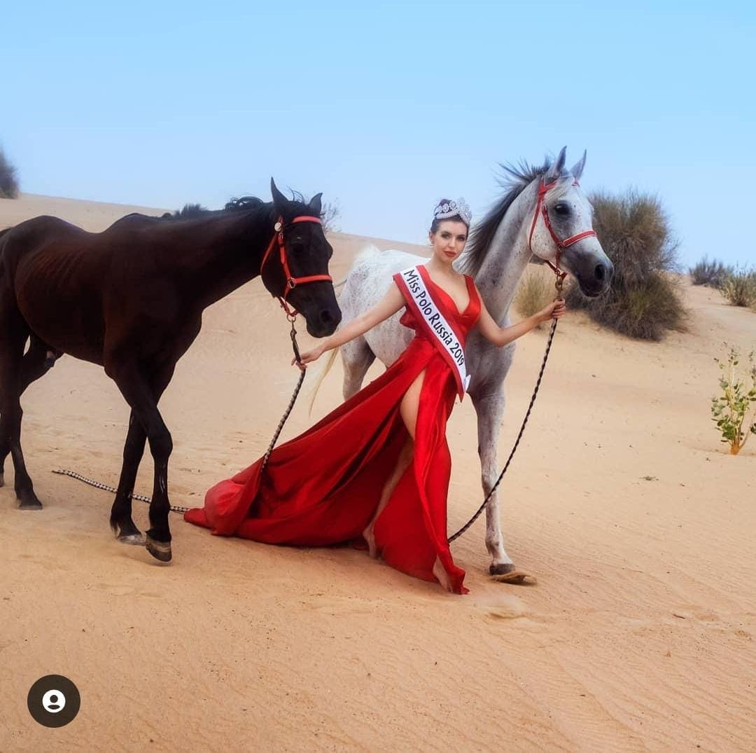 Mujer con recogido y tiara en desierto arenoso entre dos caballos, sosteniendo las riendas y vistiendo un largo vestido rojo.
