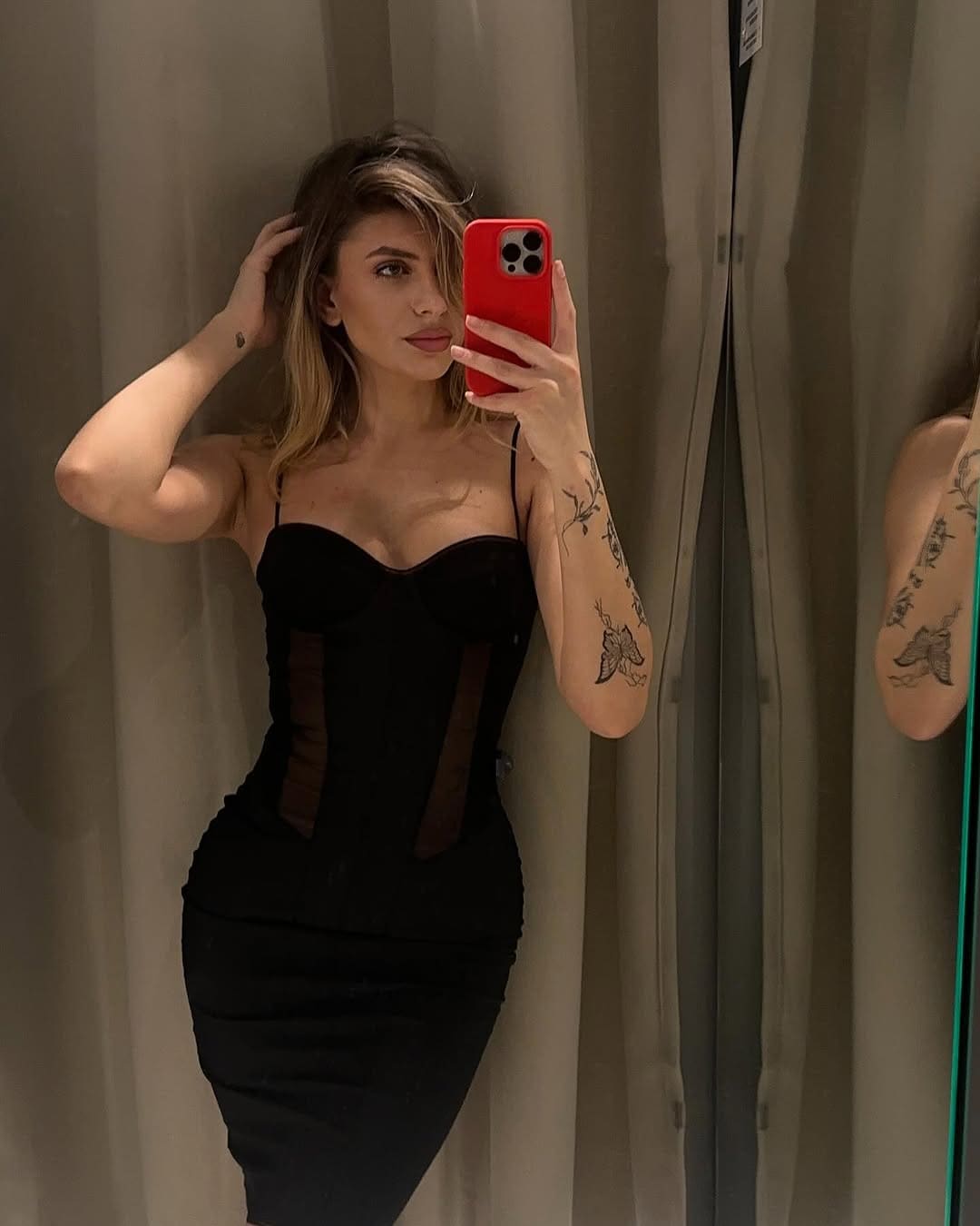 Mujer rubia tomando una selfie en el espejo con vestido negro sin tirantes y telaraña lateral.