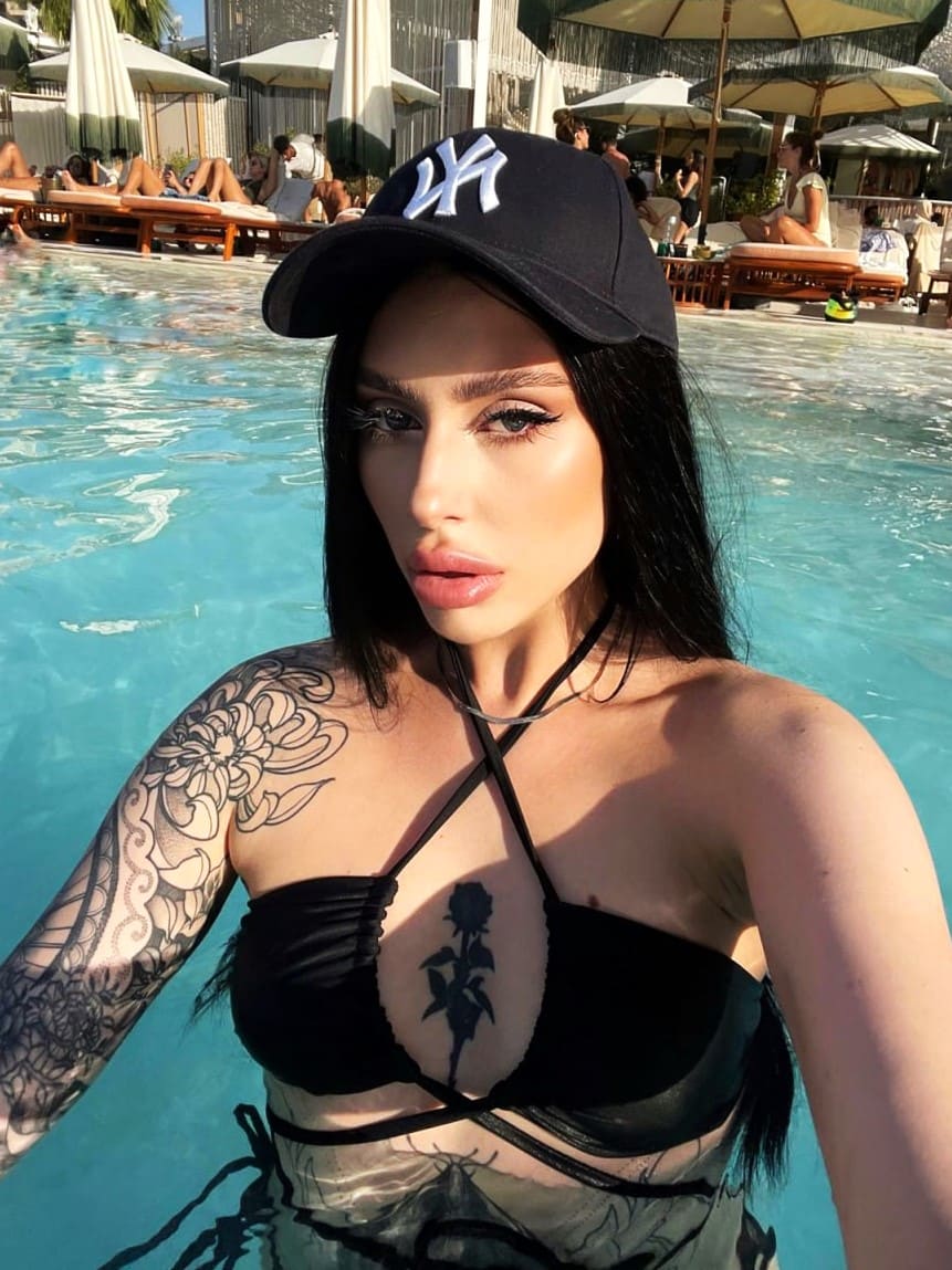 Mujer con gorra negra y top halter en una piscina, mostrando tatuajes florales en el brazo y pecho, junto a tumbonas.
