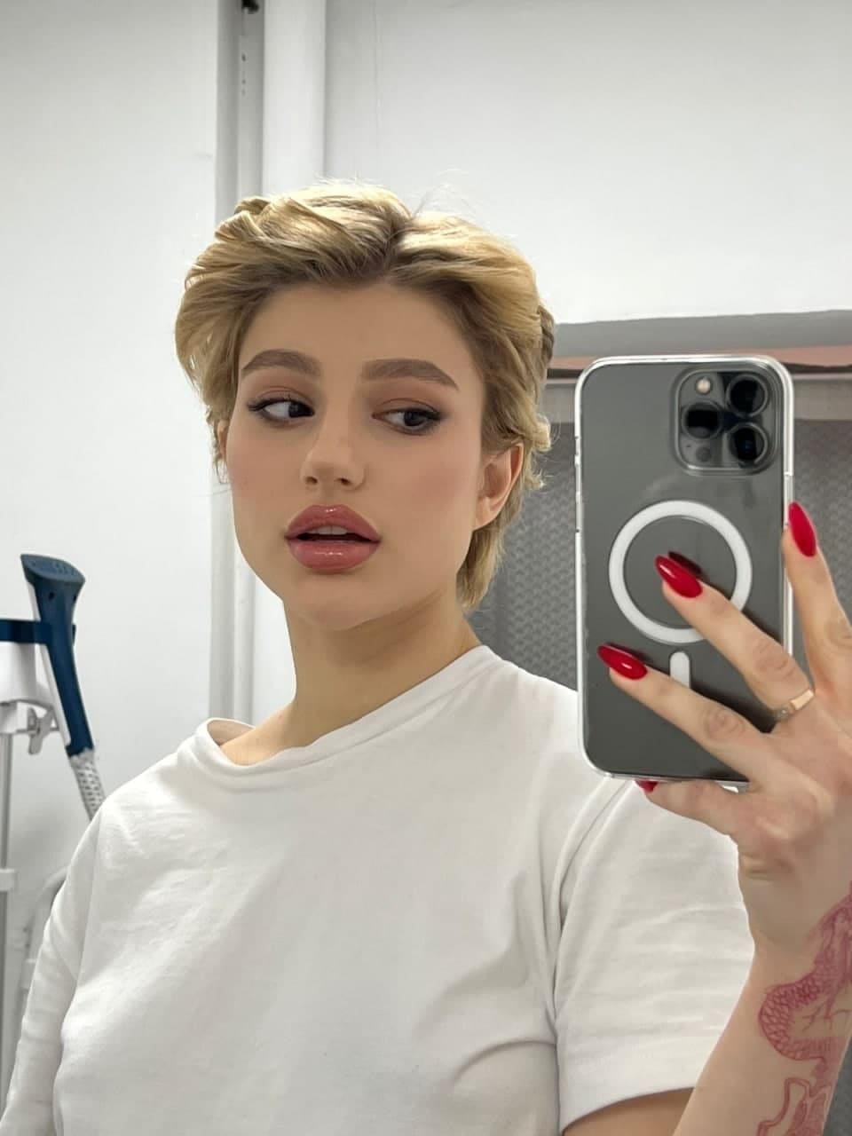 Persona con pelo rubio corto y camiseta blanca sosteniendo un teléfono gris contra una pared blanca, con esmalte de uñas rojo visible en la mano.