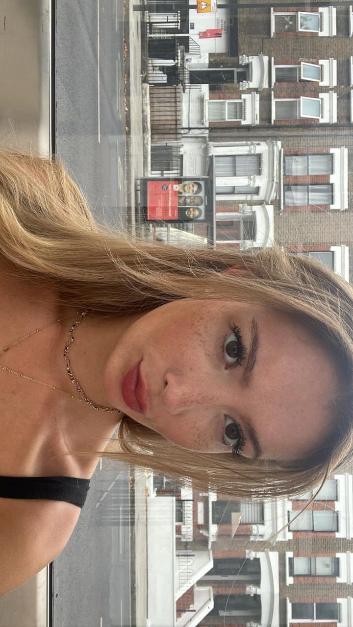 Clara leaning by a window in a black strap top with street buildings behind.