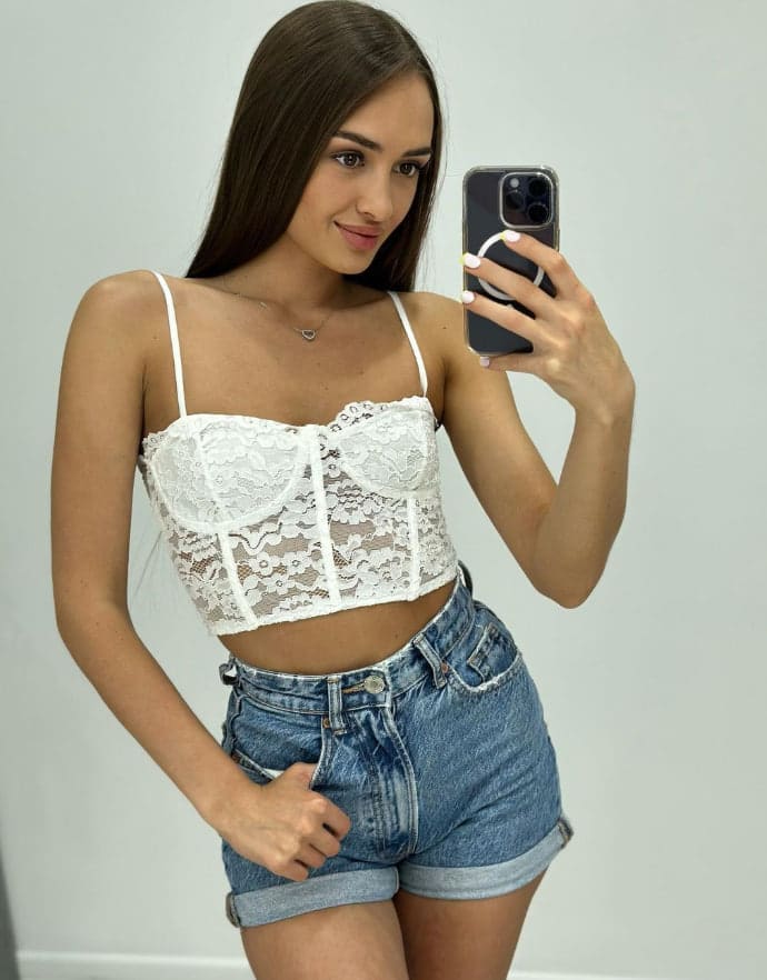 Masha si scatta un selfie allo specchio con top in pizzo bianco e shorts in denim a vita alta contro un muro chiaro.